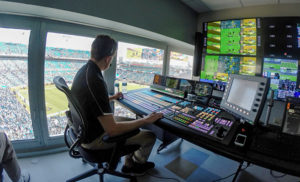 A person sits at the controls of a video display, in a room with a view from above the field at a sporting event. 