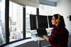 Woman sitting at a window ledge, operating computers, with a view of buildings in a downtown city scene. 