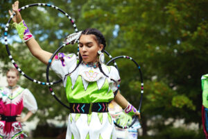 A person performs a dance outside, which incorporates large rings and traditional indigenous attire. 